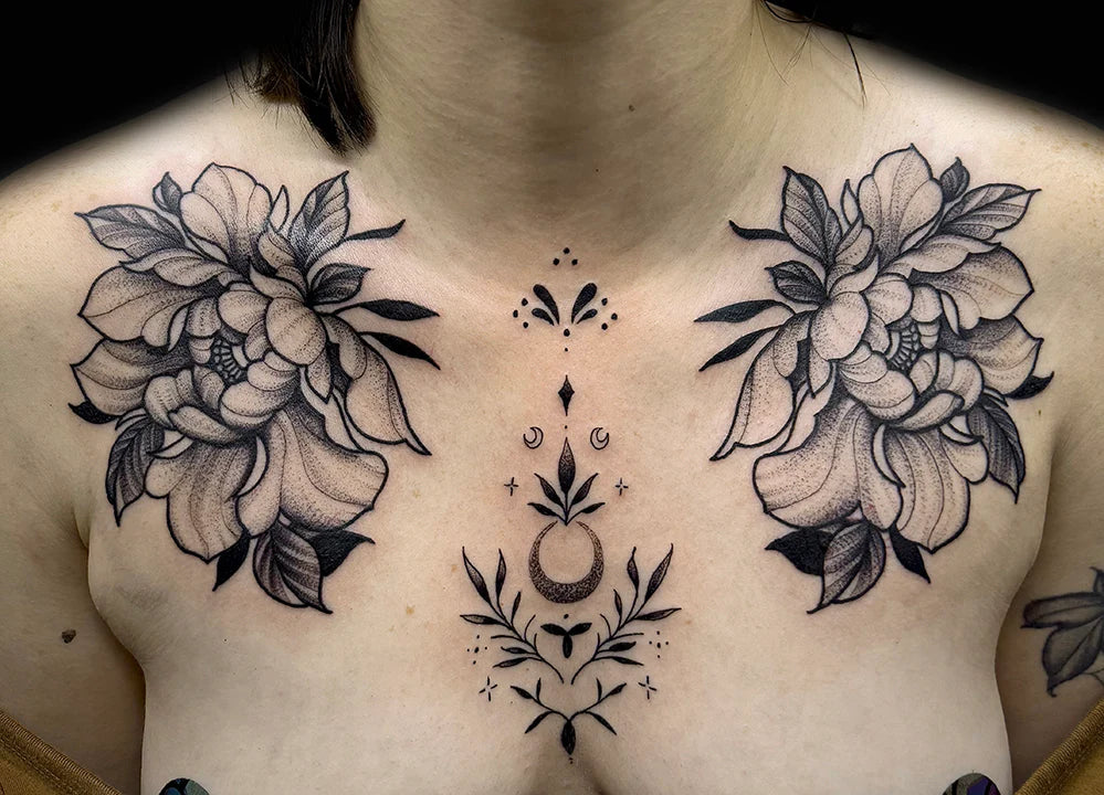 A symmetrical black and gray chest tattoo featuring two large, intricately shaded peony flowers on the collarbones, flanking a delicate fine-line ornamental design down the center.
