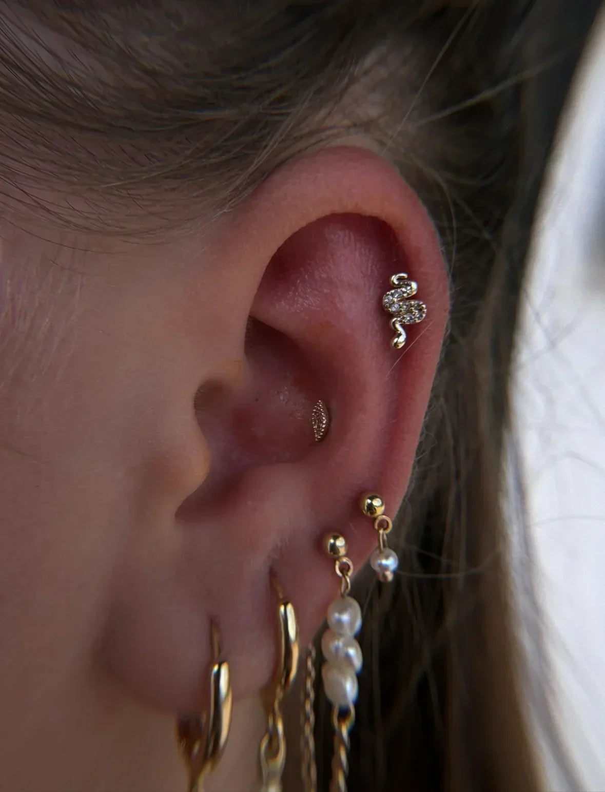 Detailed view of a fully curated ear mapping arrangement, showcasing a crystal-encrusted gold snake flat cartilage piercing, a conch stud, and multiple hanging gold and pearl lobe earrings.