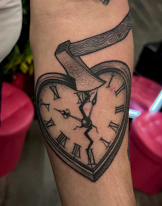 A black and grey stippled tattoo on a forearm featuring a heart-shaped clock with Roman numerals on its face. A large wood-textured axe is shown splitting down into the top of the clock, causing a prominent crack to run down the center of the clock face.