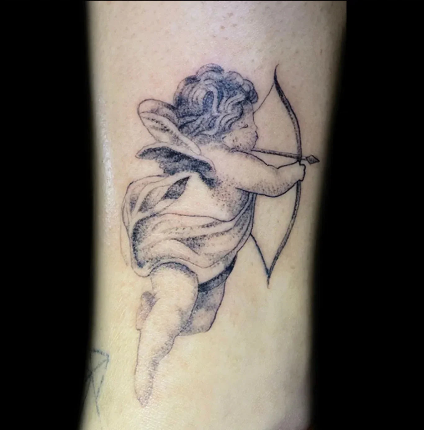 Close-up of a detailed black and grey tattoo on a limb of a small, traditional cherub with wings, drawn as if floating and aiming a bow and arrow. The tattoo has soft dot work shading.
