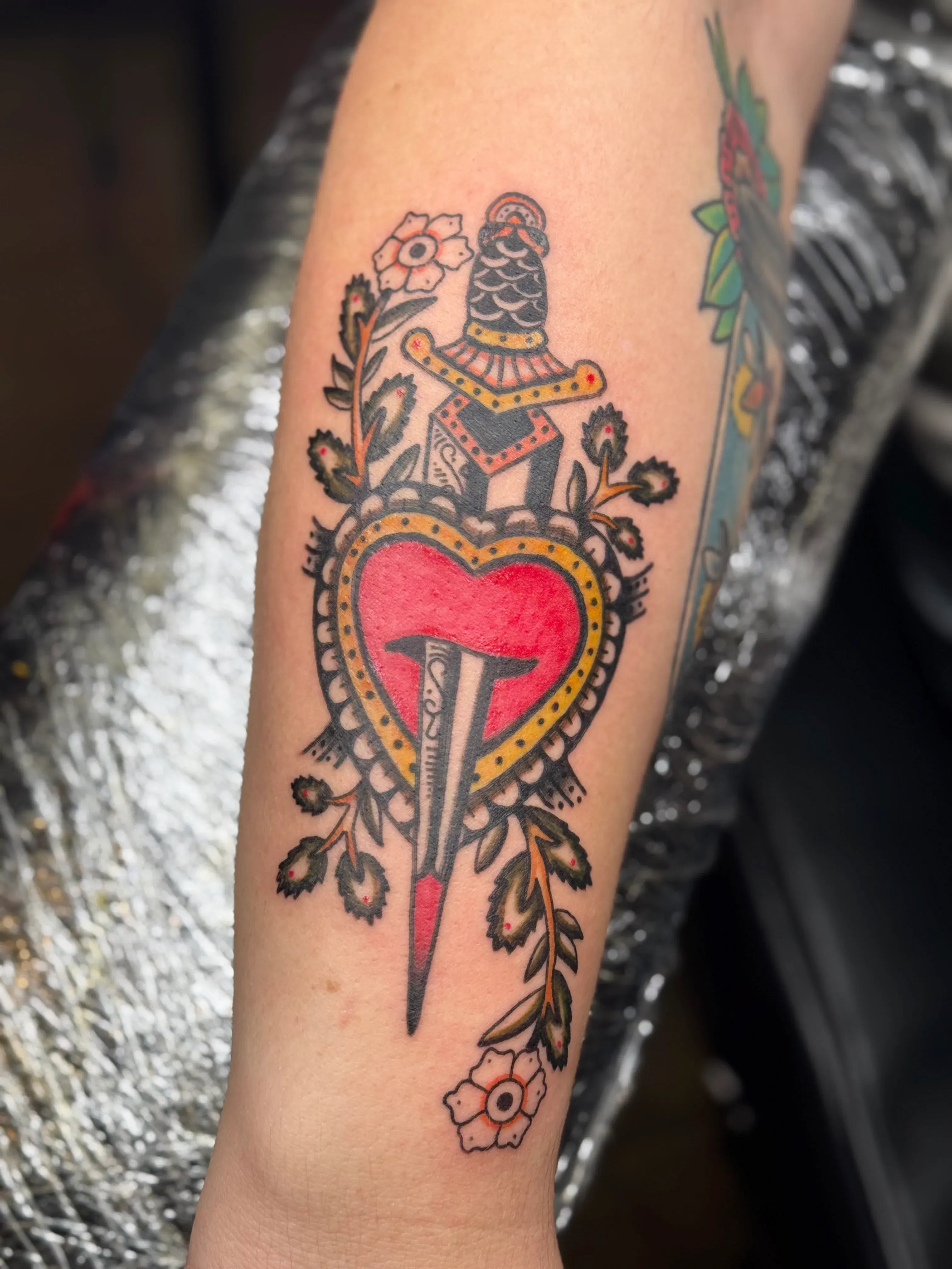 Traditional tattoo of a dagger piercing a bold red heart surrounded by leaves and flowers.