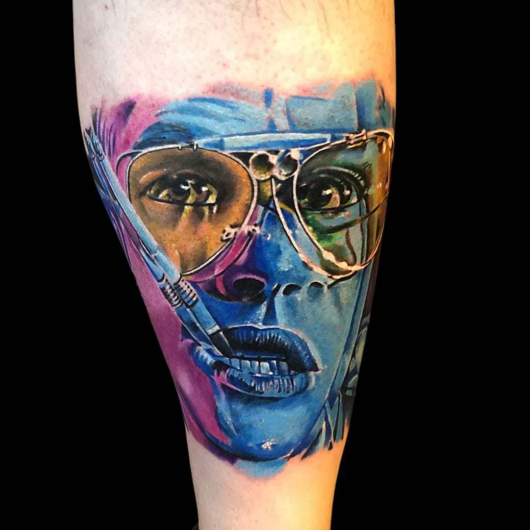 A highly detailed, colorful realism tattoo of a person wearing aviator glasses and biting a cigarette. The piece uses a bold palette of blues, purples, and yellows, creating a saturated, painterly effect on the skin.