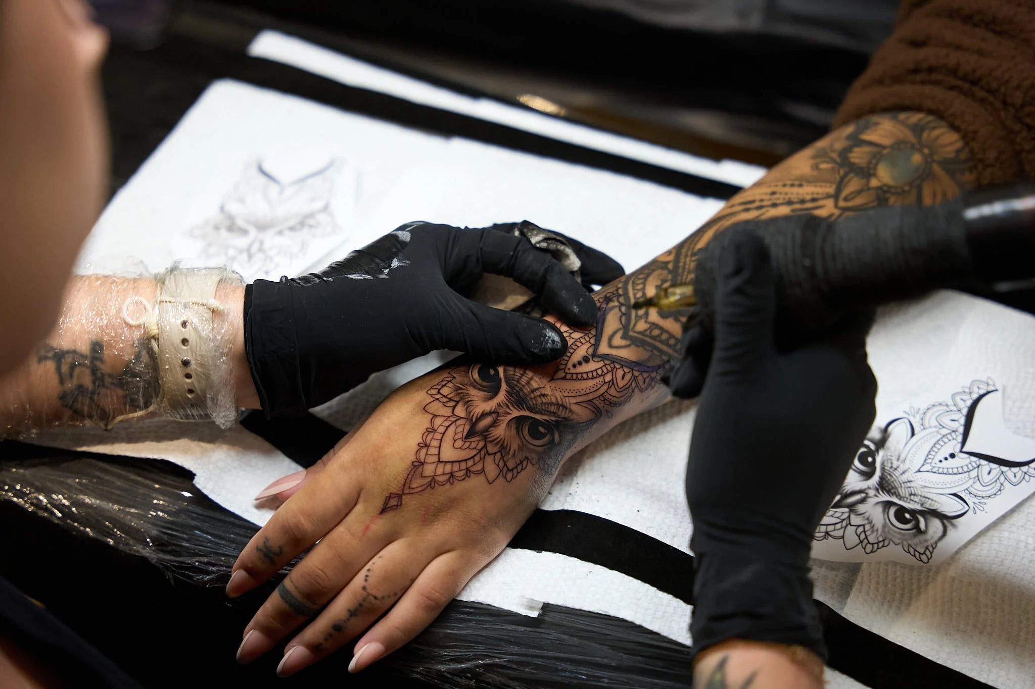An overhead close-up of a tattoo artist wearing black gloves carefully applying fine line details to a black-and-grey owl tattoo on a client's hand and wrist, with paper stencils visible on the covered workstation.