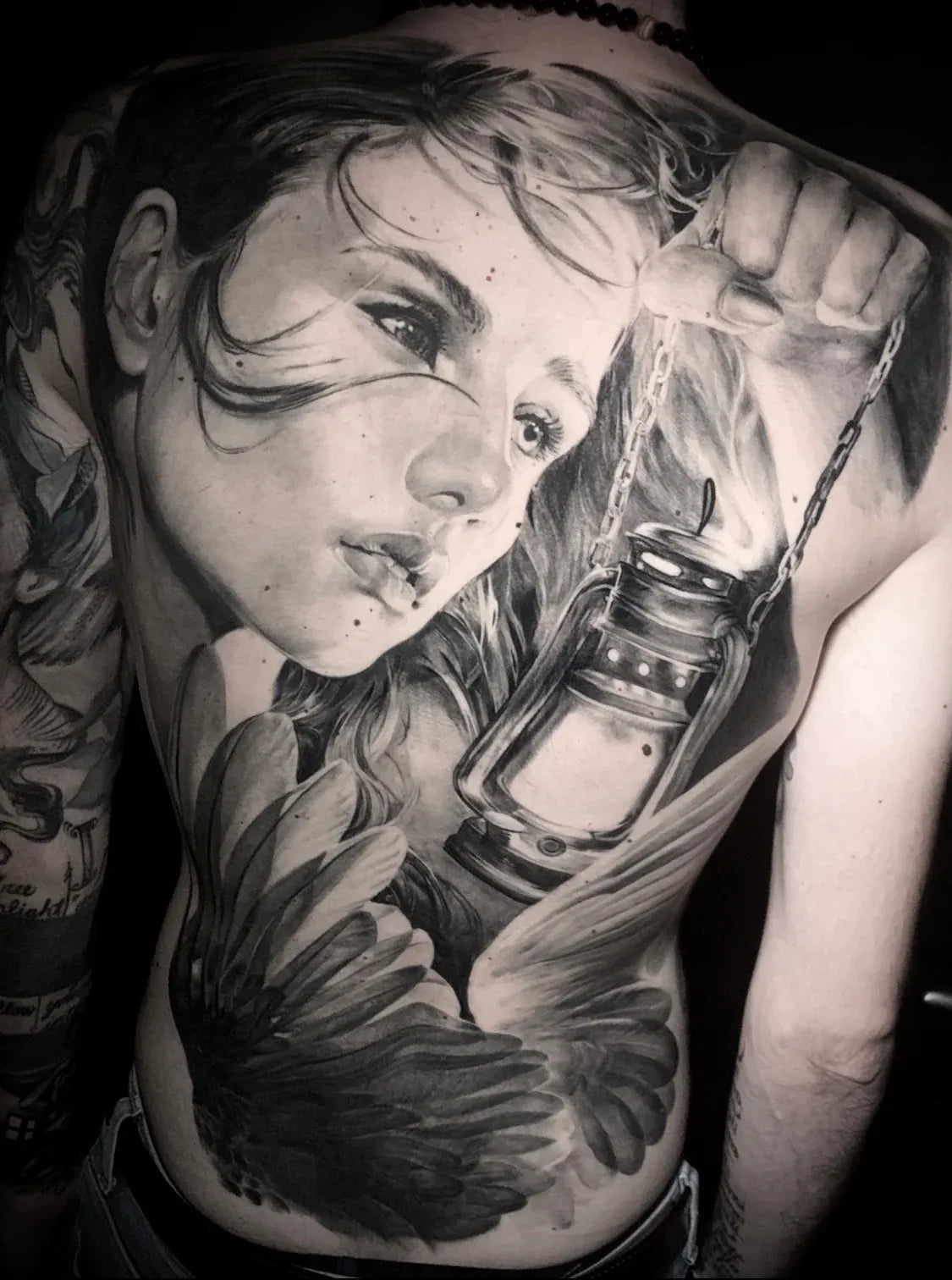 Black and grey full-back tattoo of a woman holding a lantern with feathery wings below.