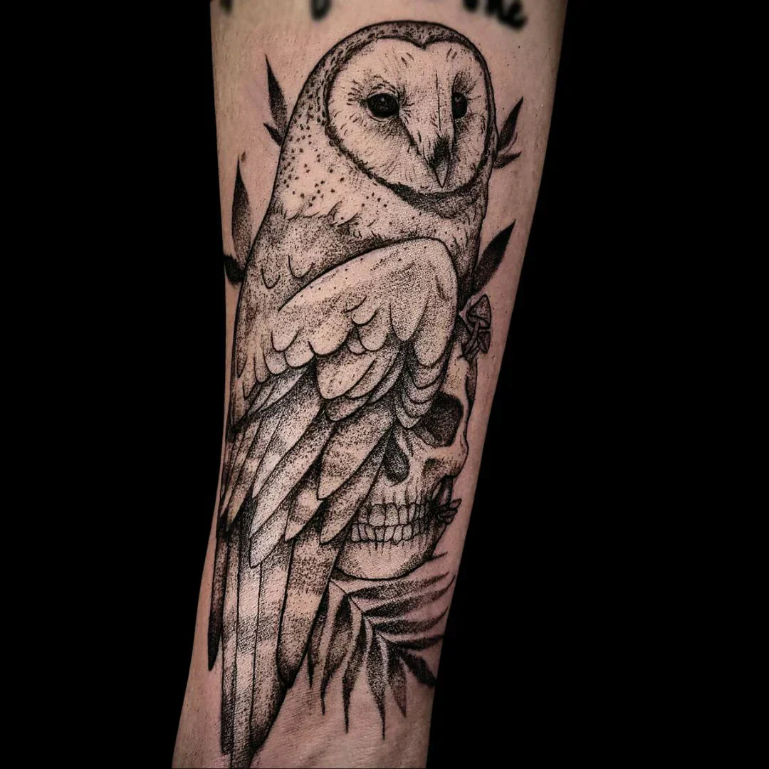 A detailed black-and-gray stipple-shaded tattoo on a forearm featuring a barn owl perched atop a human skull, surrounded by delicate leaves and small mushrooms.