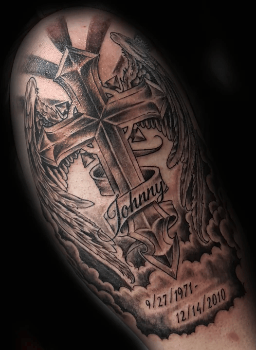 A black and gray memorial tattoo featuring a large 3D cross with angel wings, rays of light, and a ribbon reading “Johnny,” along with birth and death dates below.