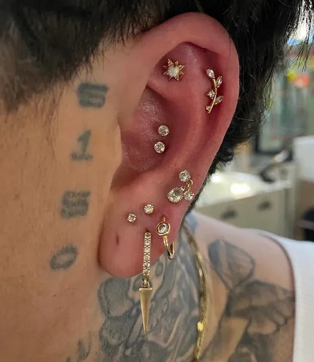 A highly curated ear display showcasing multiple gold and diamond studs along the lobe and helix, a double conch piercing with two bezel-set diamonds, and a gold leaf-clutching branch in the upper flat.