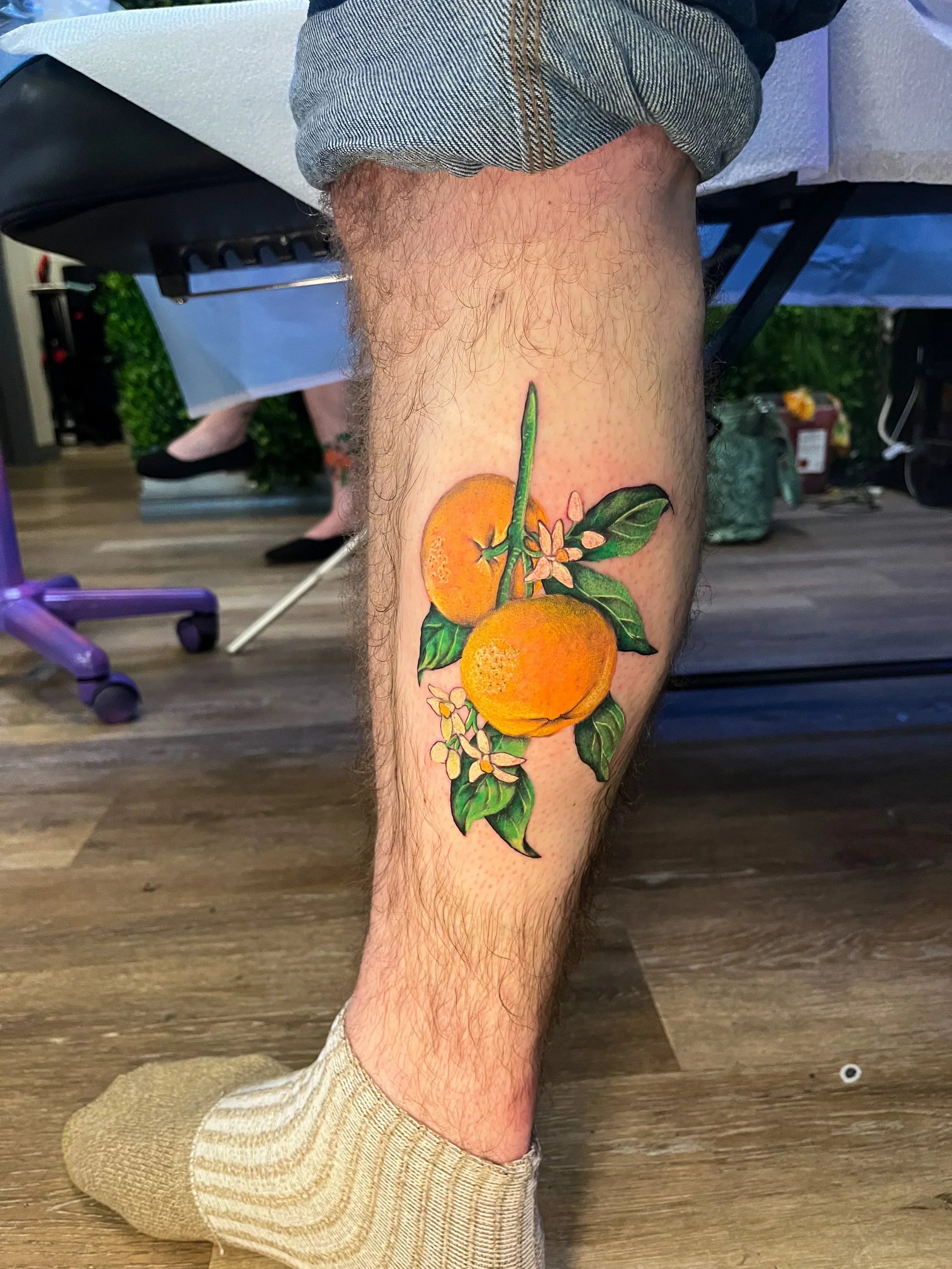 A realistic color tattoo on a person's lower leg of a branch with two bright oranges and small white citrus blossoms. The design utilizes rich orange and green tones with fine shading to capture a natural look.