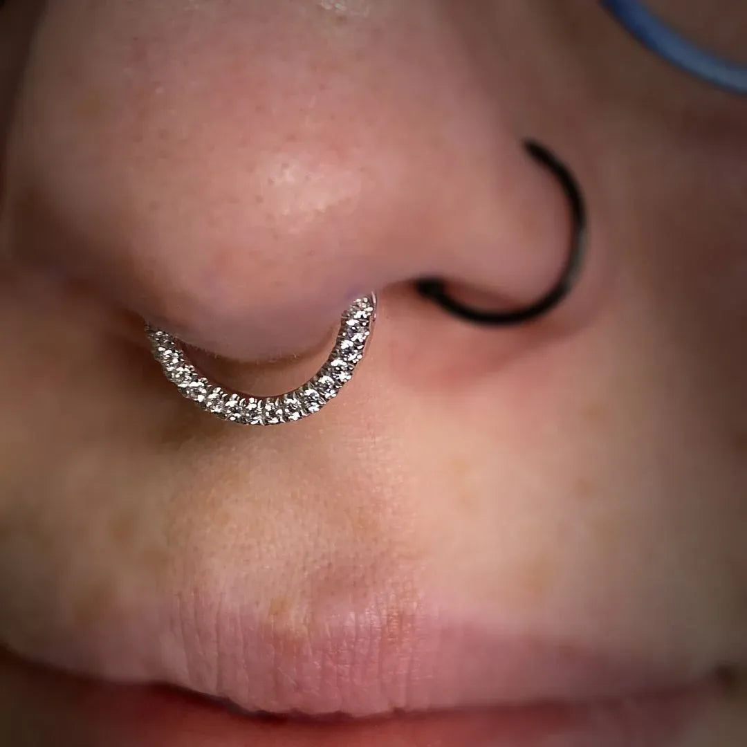 Close-up of a nose featuring a septum piercing with a sparkling, gem-encrusted hoop and a simple black ring in the nostril.