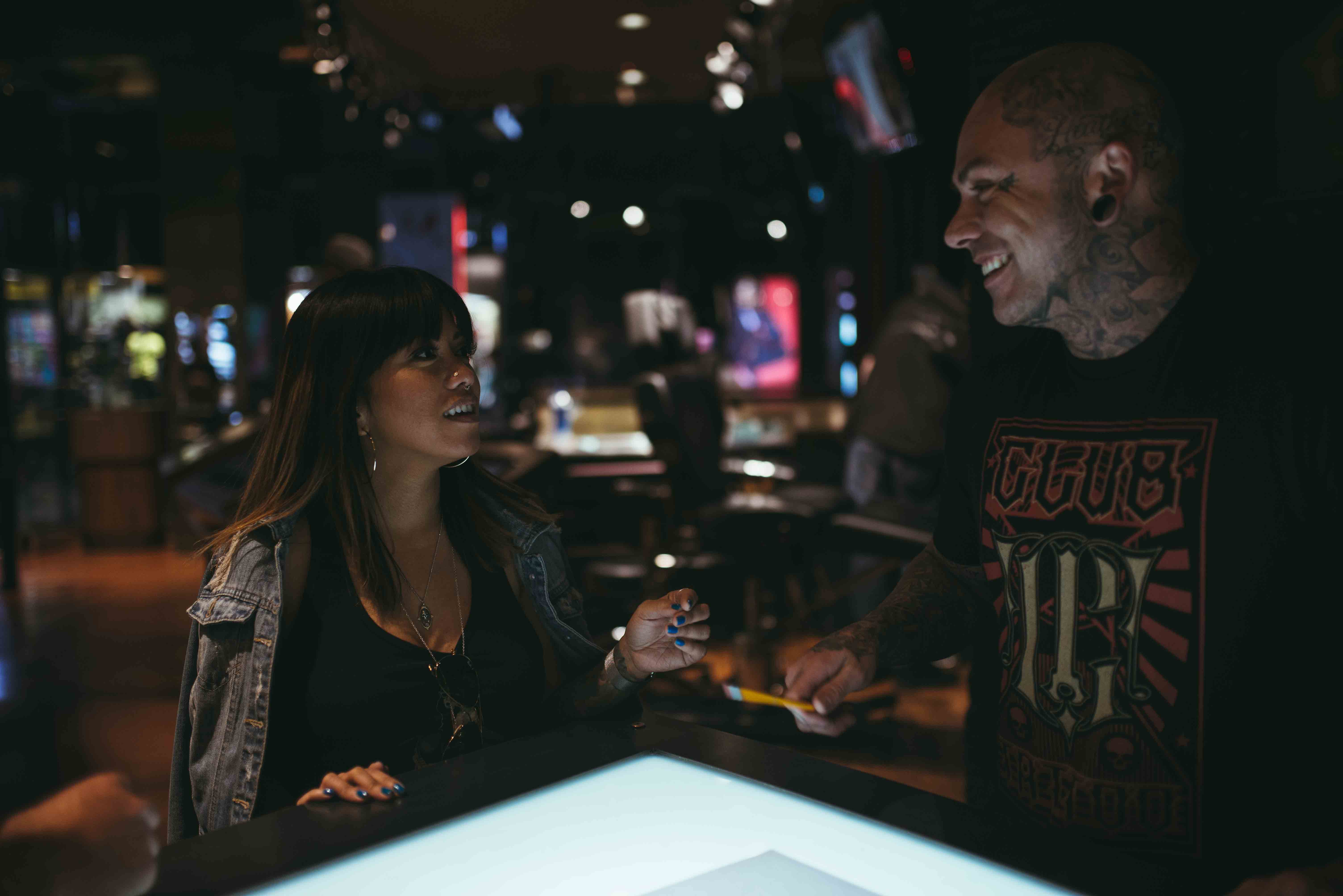 A heavily tattooed Club Tattoo staff member in a black branded t-shirt smiling and conversing with a female client as they collaborate over a large glowing digital design table in a high-end, warmly lit studio environment.
