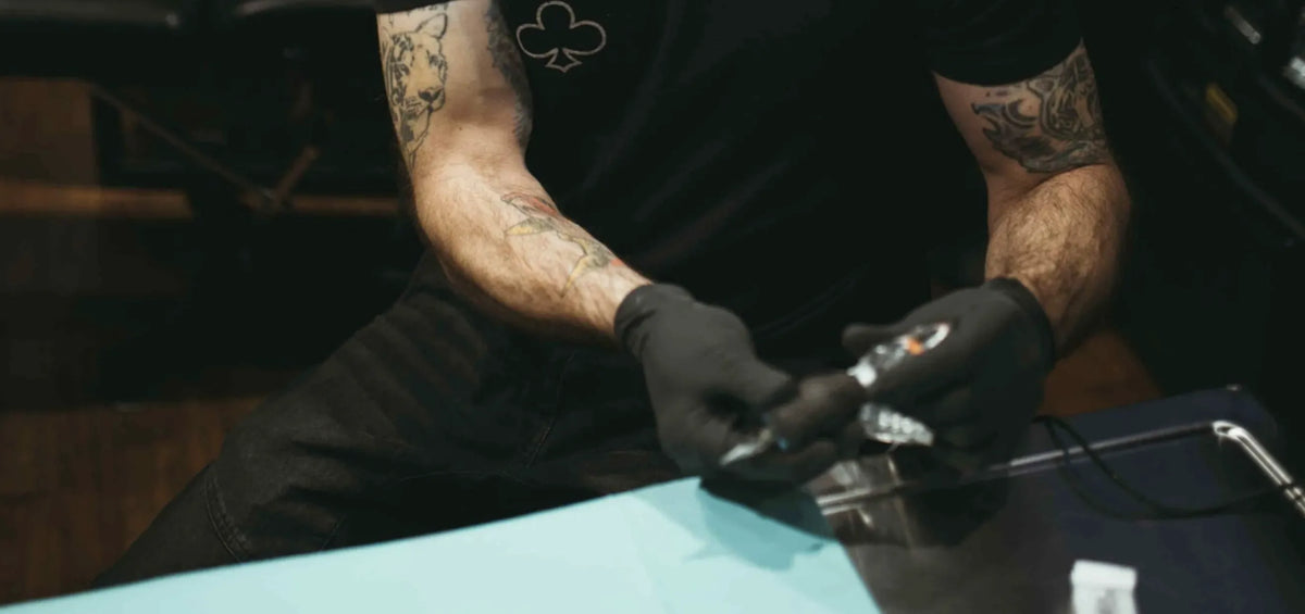 A tattoo artist wearing black gloves and a Club Tattoo shirt prepares a sterile tattoo machine at a workstation.