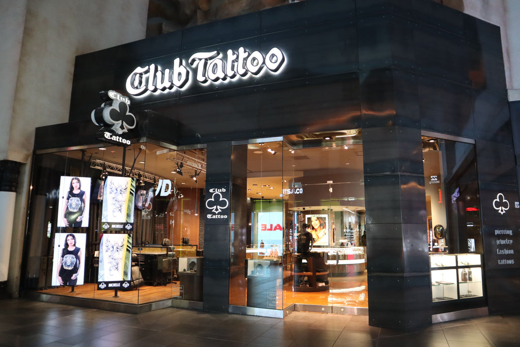 Exterior view of the Club Tattoo studio featuring large illuminated signage, window displays with tattoo artwork, and a modern black metal facade.