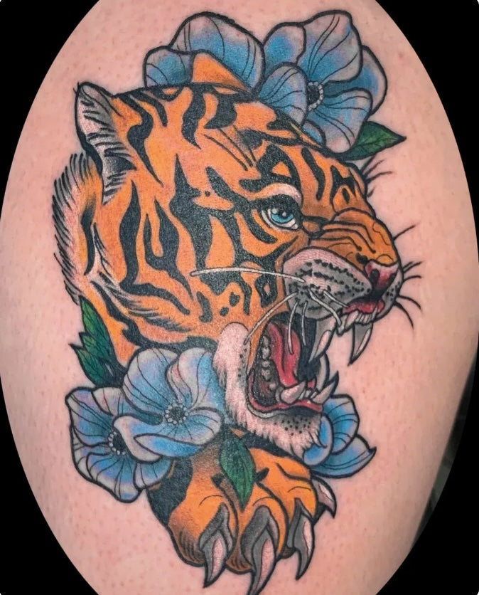 Neo-traditional tiger head tattoo with bold black stripes, bright orange fur, and vivid blue flowers surrounding it, showing the tiger mid-roar with open jaws and sharp teeth